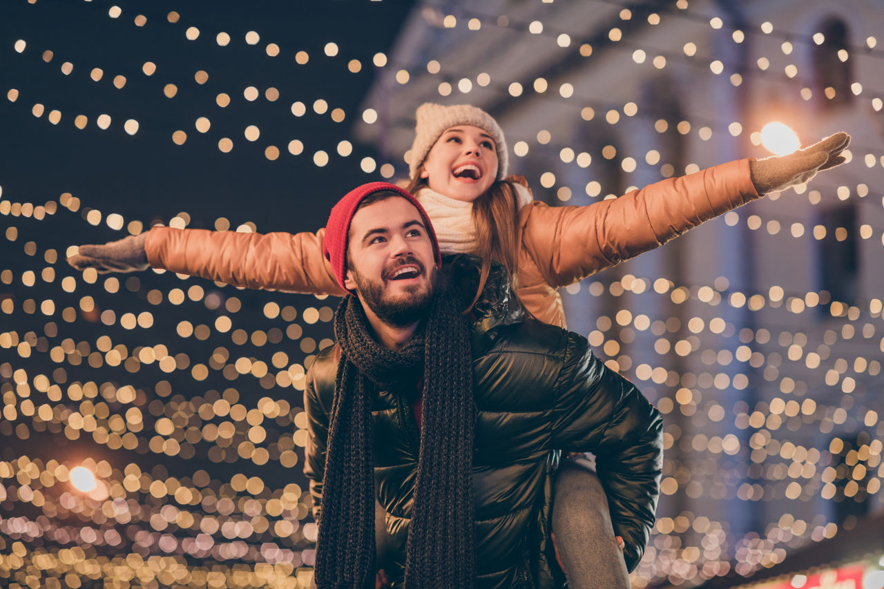 Photo of positive couple having fun christmas x-mas around evening, outside illumination boyfriend piggyback girlfriend holding hands fly